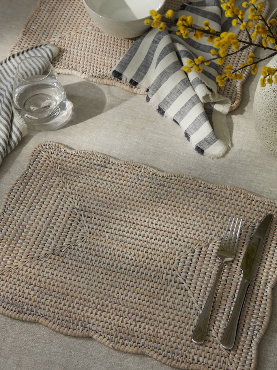 Ceres white rattan scalloped placemat styled with striped napkin, glass and yellow flowers, neutral spring table setting woven placemat UK home decor