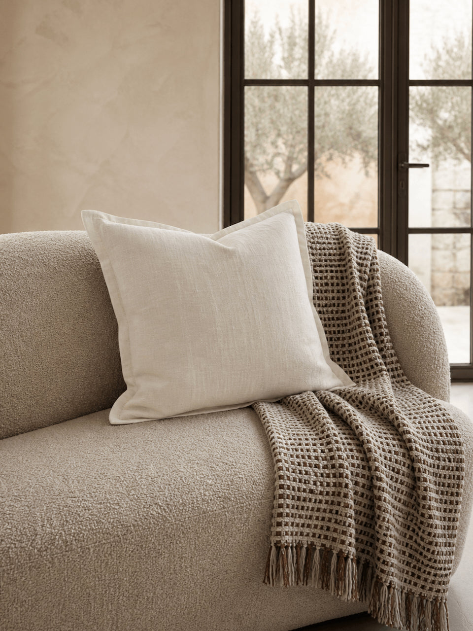 Luxury cream cotton linen cushion cover styled on a textured boucle sofa with a woven neutral throw in a warm contemporary interior.