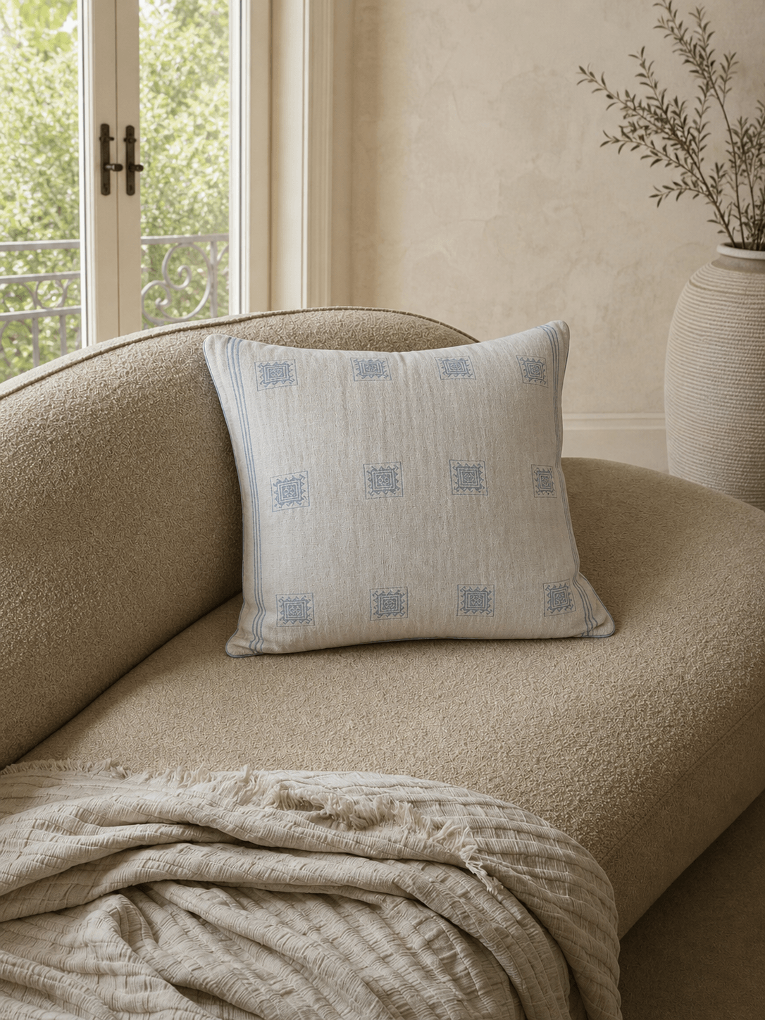Luxury cream and blue patterned BCI cotton cushion cover with woven geometric detailing and soft blue piping, styled on a textured boucle armchair in a warm contemporary interior.