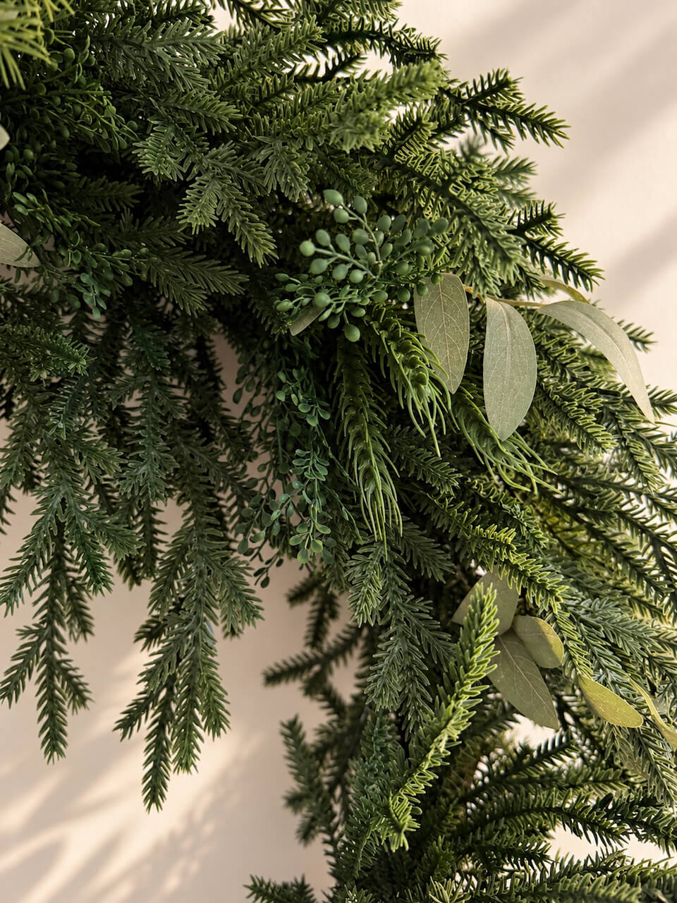 An artificial green wreath made of pine and fir branches with small green berries and eucalyptus leaves, shown against a warm beige background.
