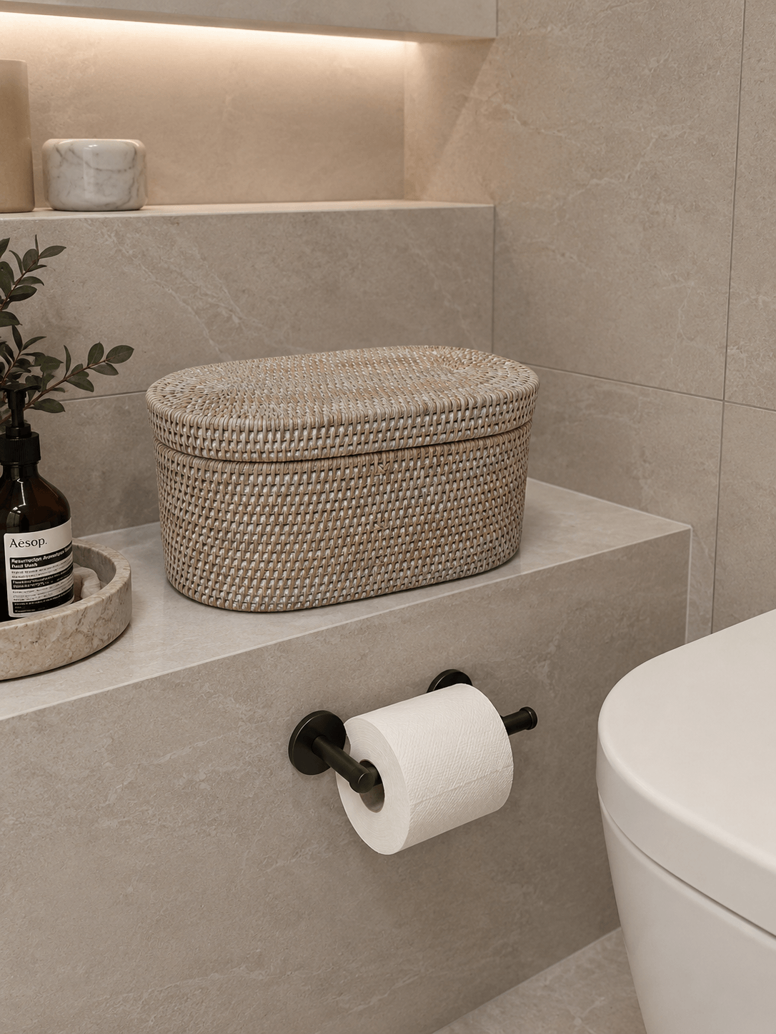 Bathroom setting with a Jarra rattan basket, toilet paper roll, and plant on a shelf.