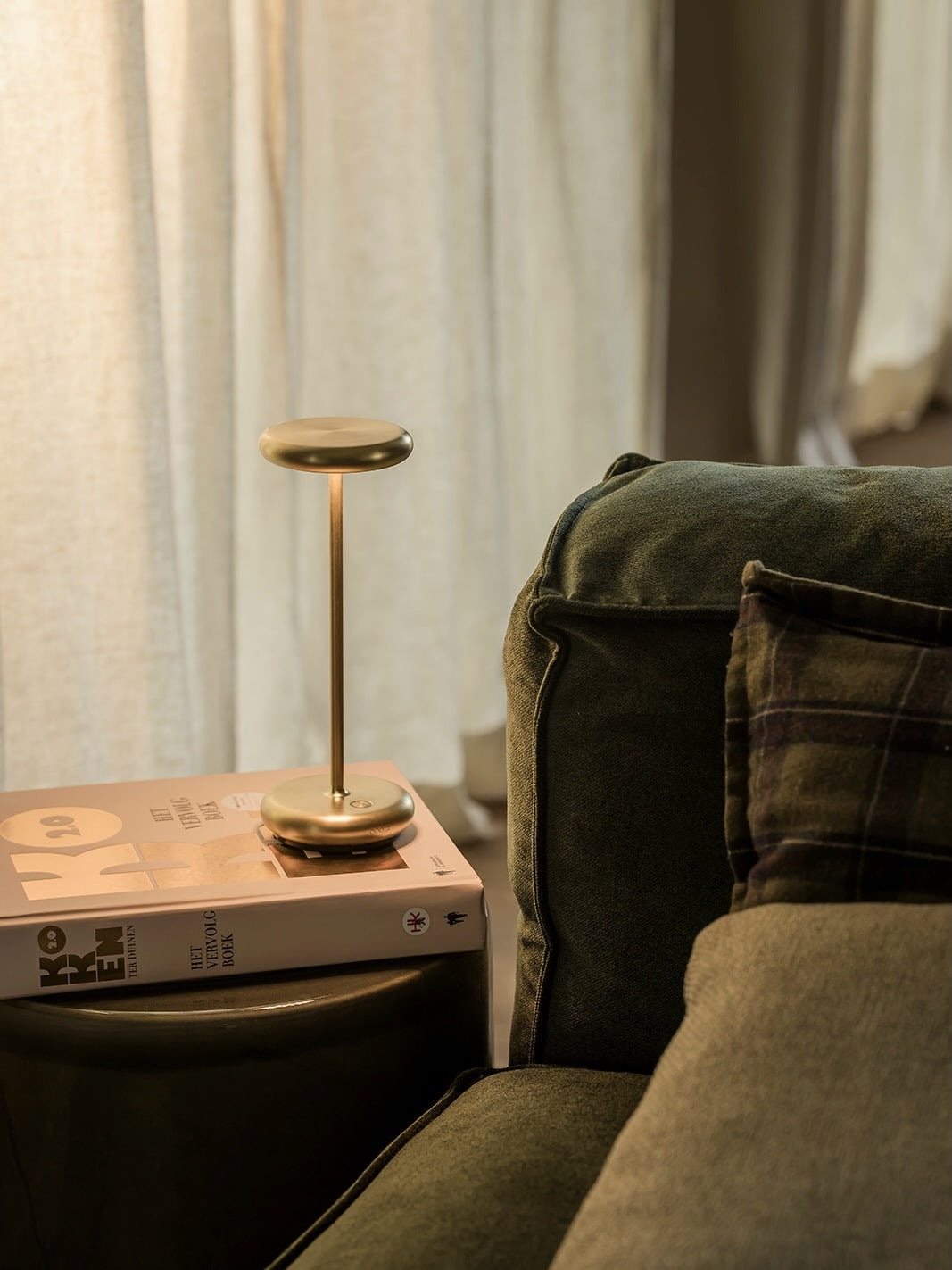 A slim brass table lamp with a circular shade and base sits on top of a book next to a dark green velvet sofa in a softly lit room.