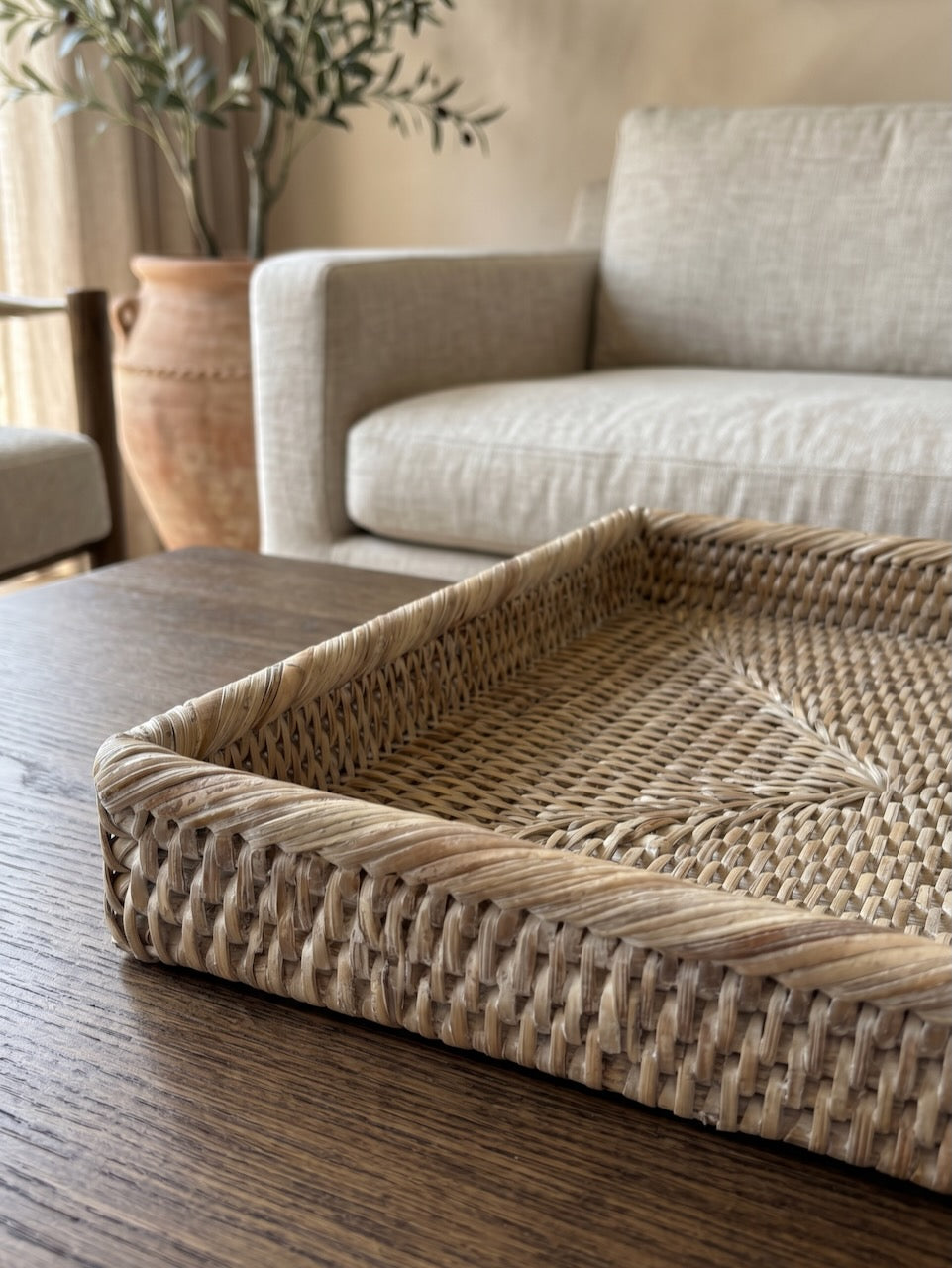 Close-up detail of a handwoven natural rattan tray on a dark wooden coffee table, showcasing the intricate weave and softly whitewashed finish in a warm neutral living room.