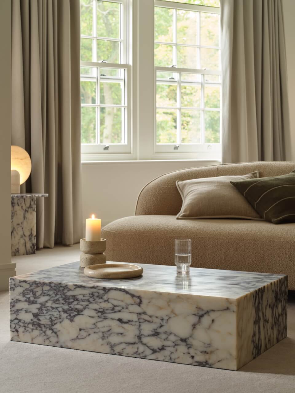 A modern low-profile rectangular marble coffee table in a room with a large window, a beige sofa, and a lamp.