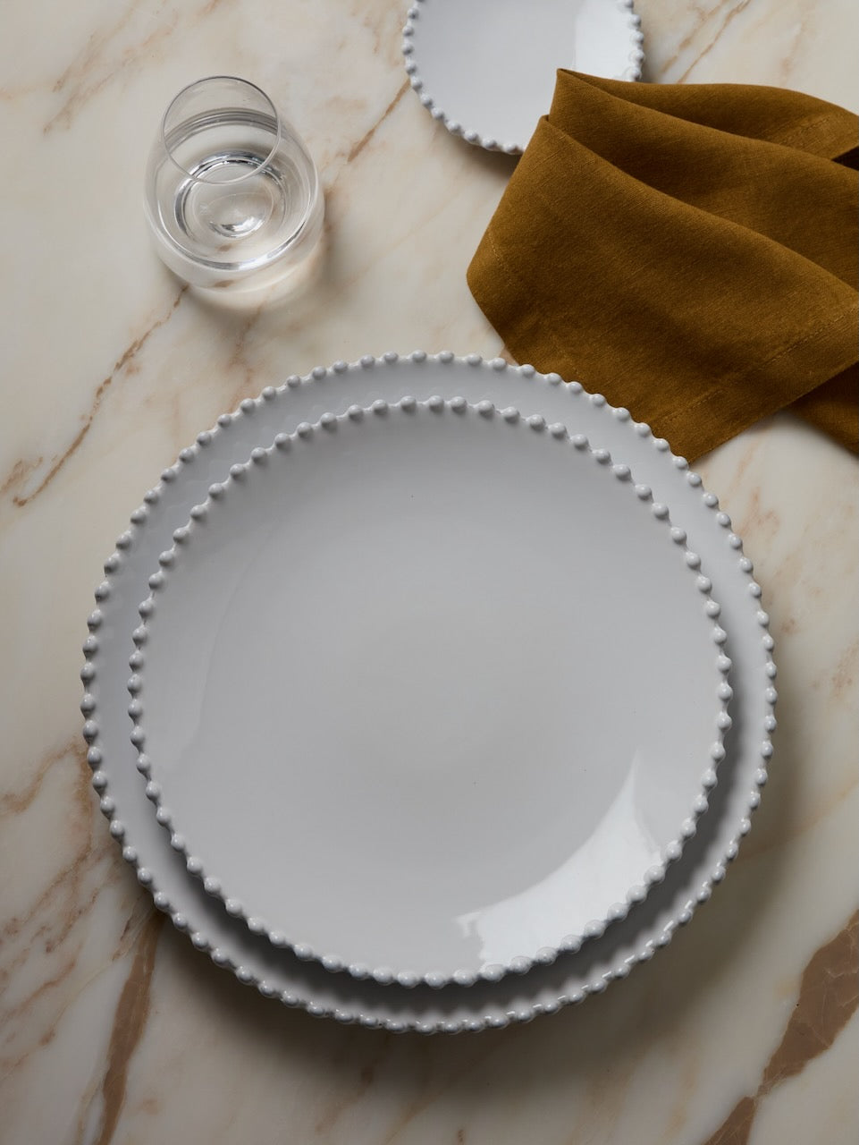 White Perl ceramic plate with decorative rim on a marble surface with a glass of water and bronze napkin.