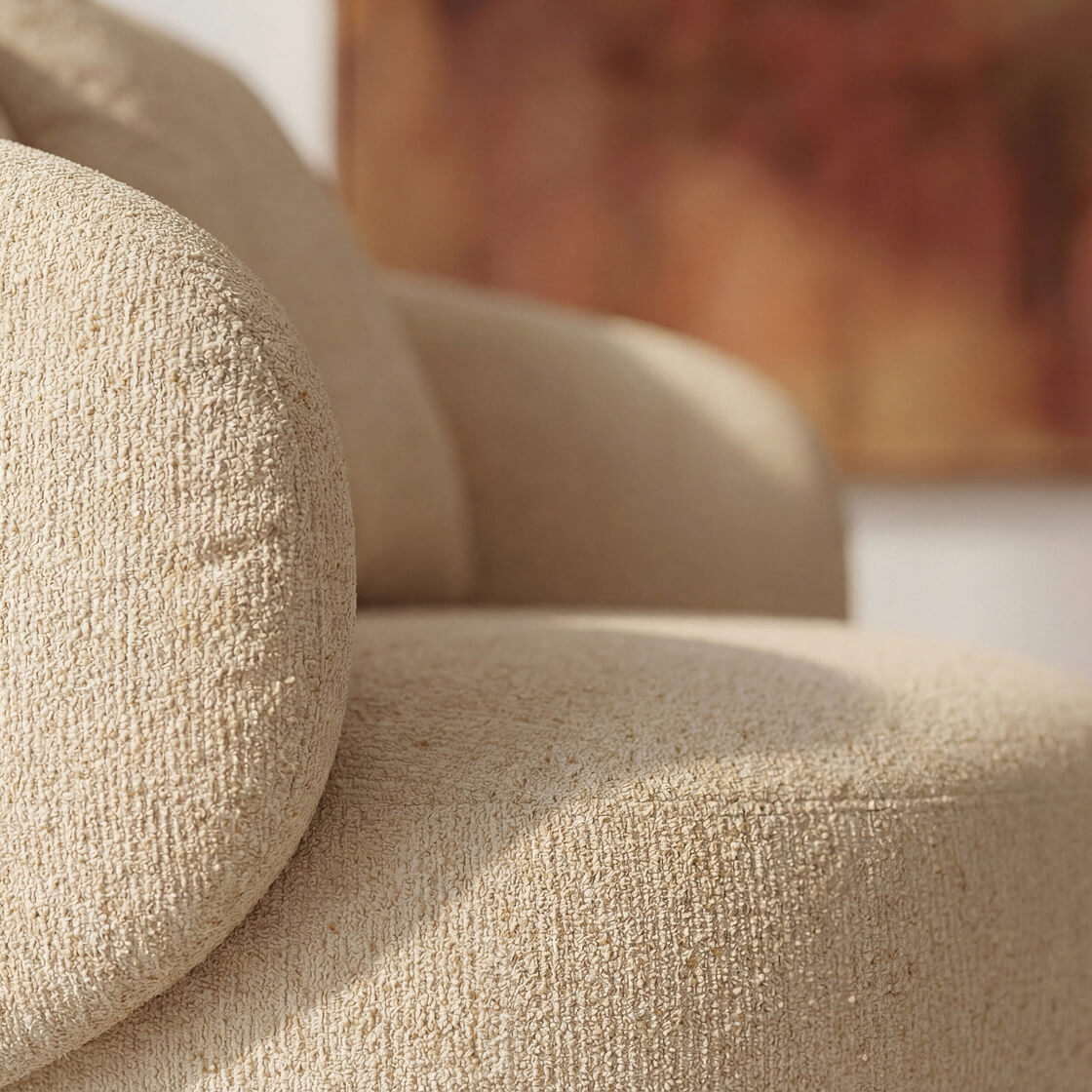 Close-up of a beige upholstered chair or sofa with a textured bouclé fabric, showing the curved armrest and seat in warm natural light.