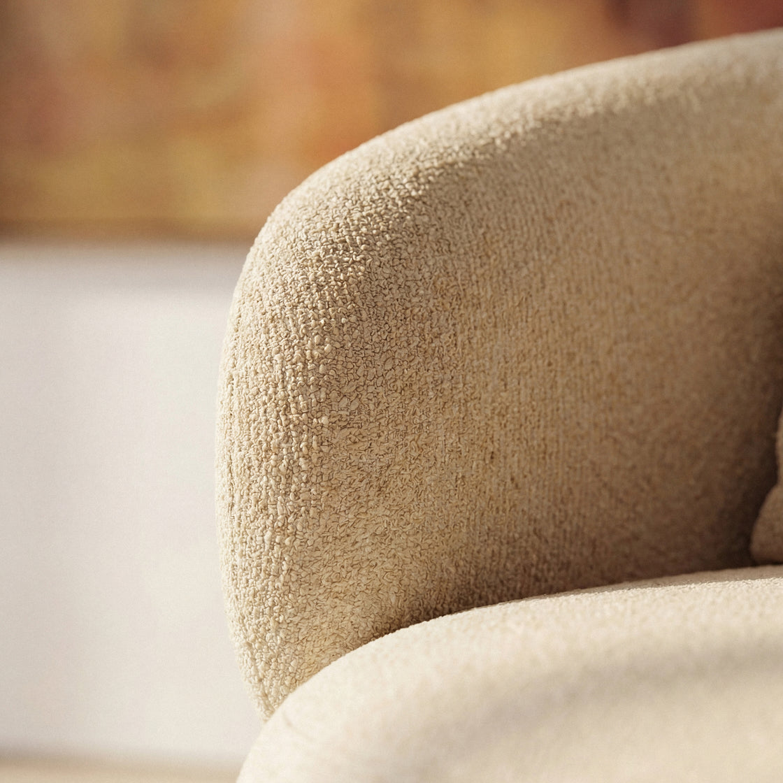 Close-up of a beige textured boucle fabric armrest on a modern upholstered chair, with a softly blurred warm-toned interior background.