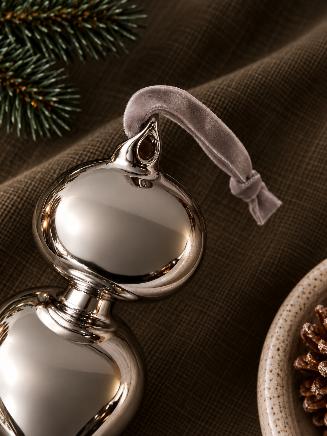 A close-up of a polished silver Christmas ornament shaped like a gourd, with a soft taupe velvet hanging loop, resting on a textured brown fabric surface with pine needles and a ceramic dish in the background.