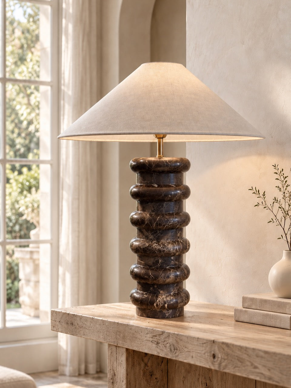 A table lamp with a dark brown marble stone column base and a beige fabric conical shade, styled on a rustic wooden table in a warm, sunlit room.