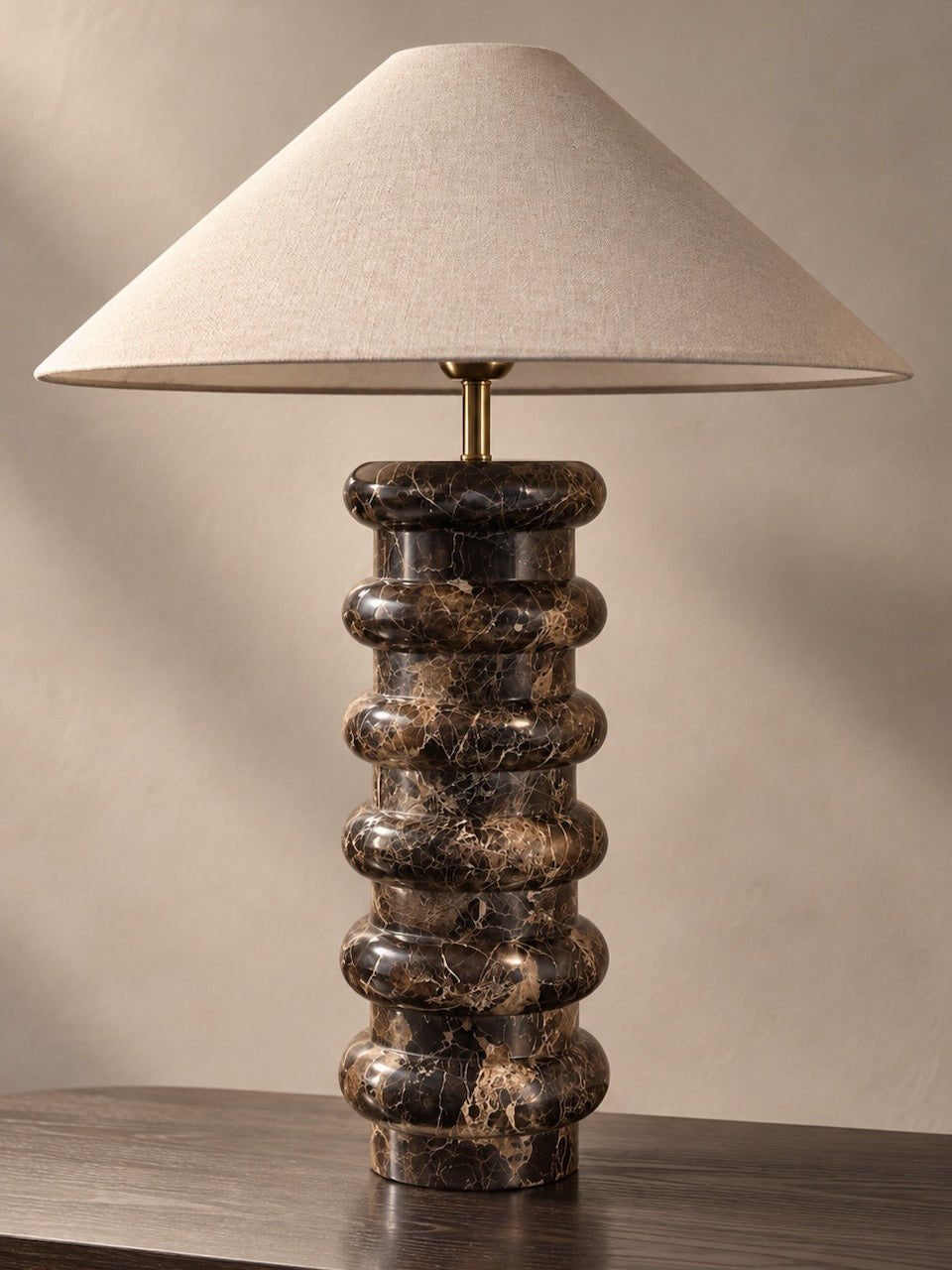A brown marble table lamp with a tapered beige fabric shade and a sculptural stacked marble base in dark brown and black with gold veining, set on a wooden surface against a neutral beige wall.