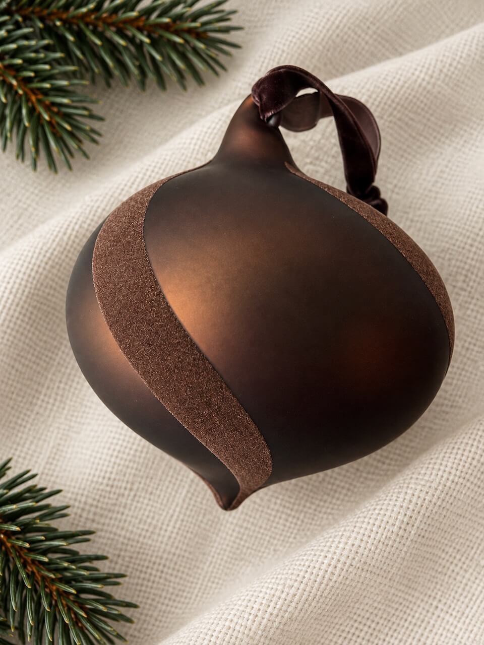 A dark brown glass Christmas ornament with a textured velvet bronze band and a dark brown ribbon hanger, styled on a cream linen fabric with pine branches in the corners.