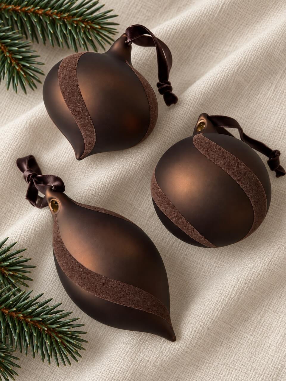 Three dark brown Christmas ornaments with textured velvet bands, arranged on a beige linen fabric with pine branches in the corners.