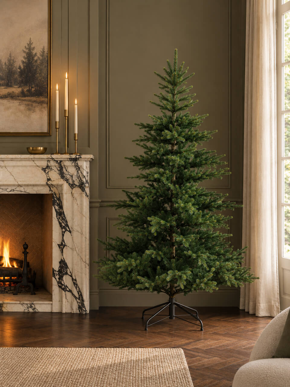 A tall artificial Christmas tree with dense green needles stands on a black metal stand in a styled living room with a fireplace, candles, wood paneling, and a window with curtains.