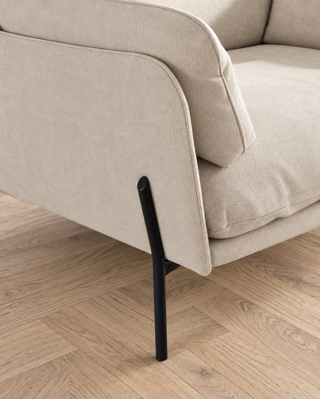 Close-up of a light beige upholstered sofa corner with a black metal leg, set on a light wood herringbone floor.