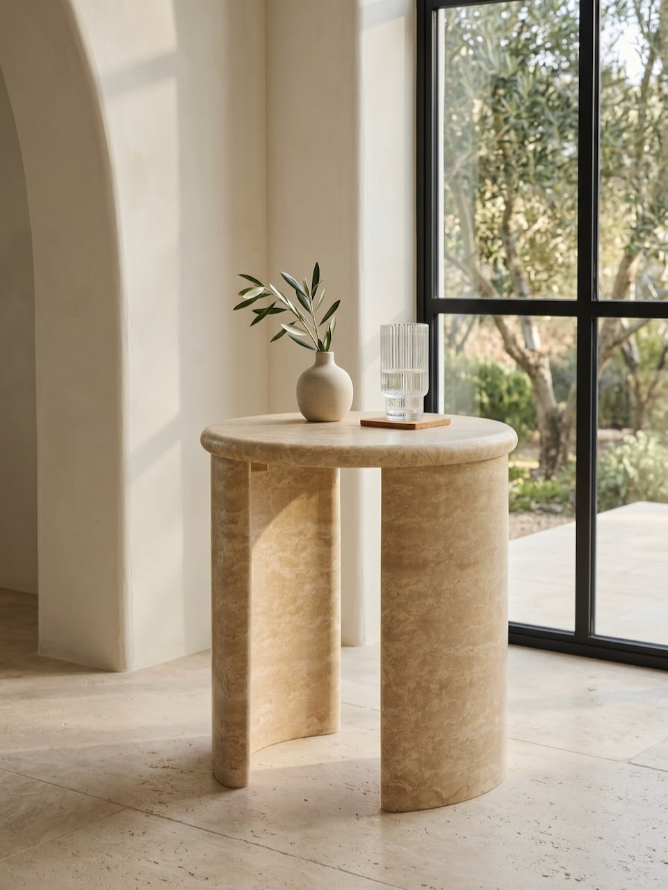 Beige travertine side table with decorative items in a bright room with large windows.