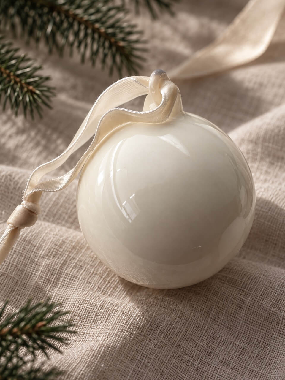 A glossy white ceramic Christmas ornament with a cream ribbon loop, resting on a textured beige linen fabric with pine branches in the background.