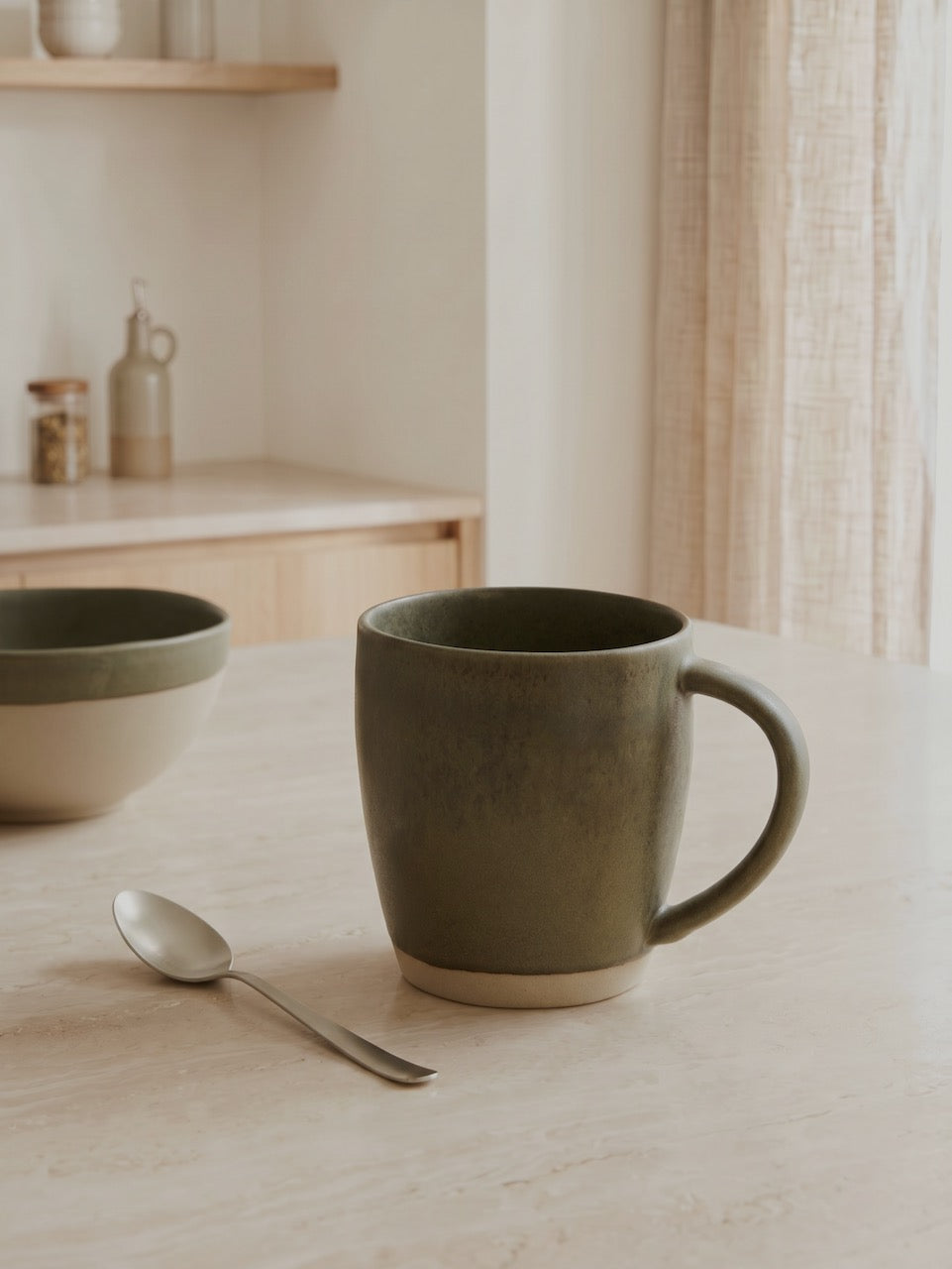 Green ceramic mug on a wooden table with a spoon and bowl in a minimalistic setting.