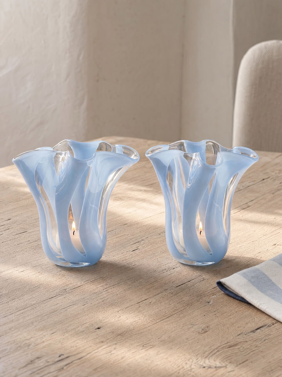 Two light blue fluted glass candle holders with a wavy, ribbed design, shown on a light wood table in a softly lit interior setting.