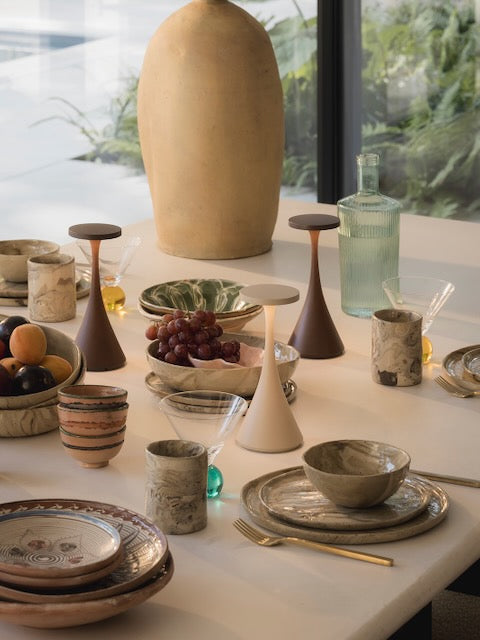 A styled dining table display featuring a collection of ceramic tableware, including plates, bowls, cups, and a central large vase, alongside glassware and wooden candlesticks in a bright, modern interior setting.