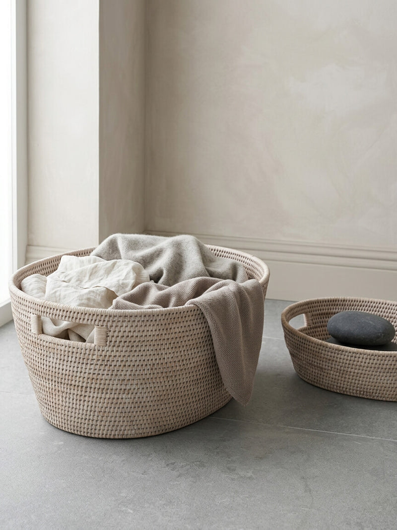 Pair of pale natural rattan storage baskets with cut-out handles, styled with neutral textiles in a modern warm-toned interior.