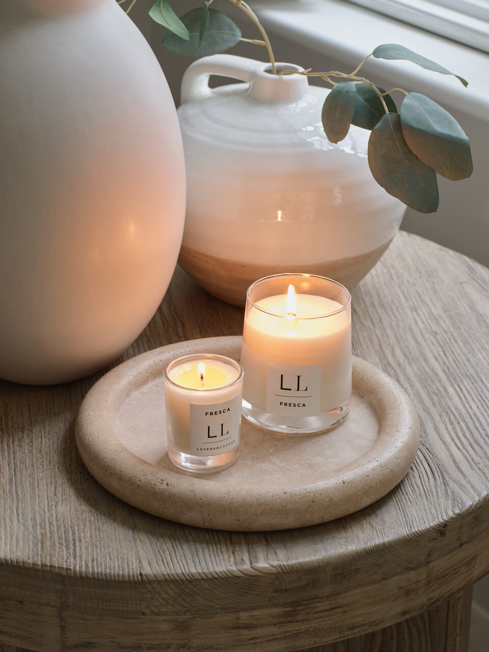 Two candles are lit, one larger and one smaller, both with white labels. They are placed on a round, beige surface with a larger white vase in the background, containing a green plant.