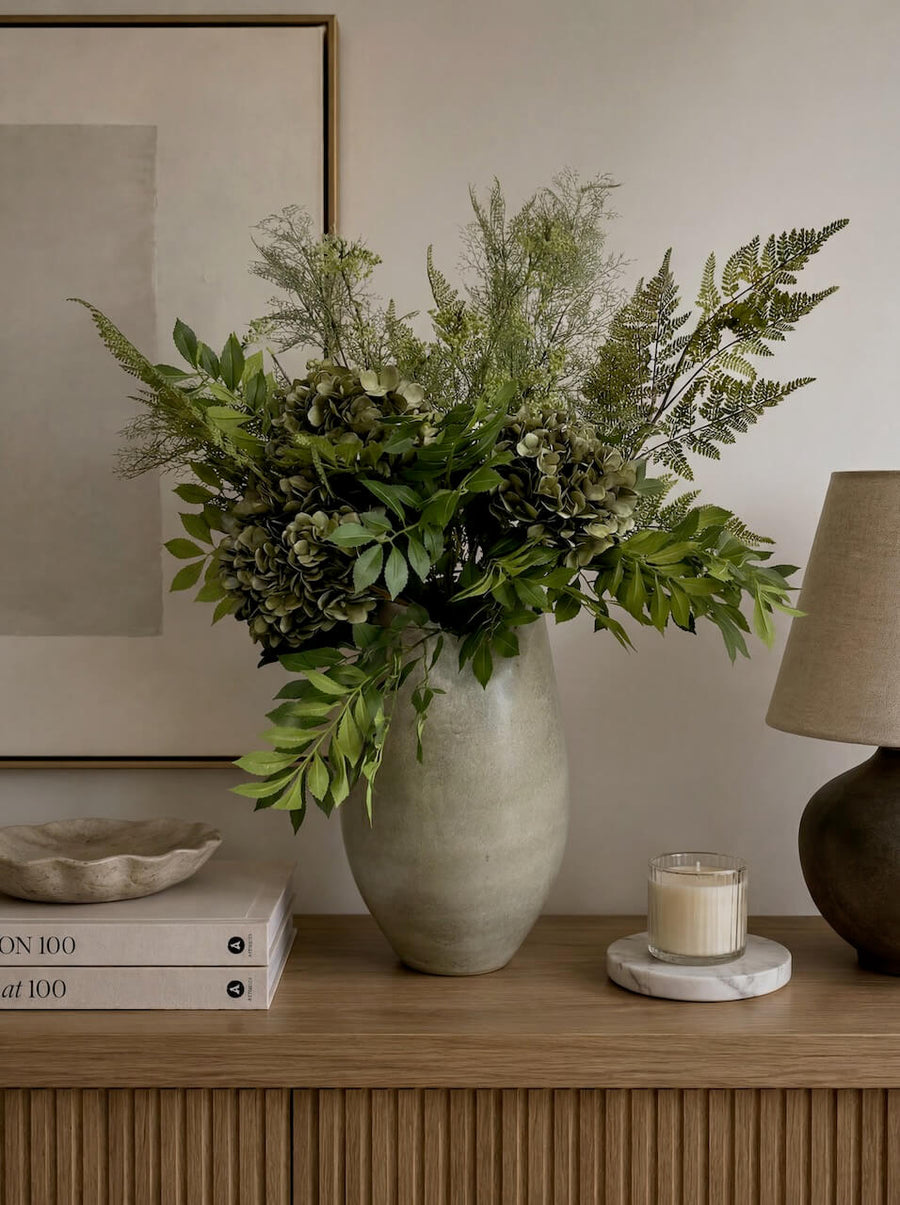 Vase with greenery on a wooden surface with books and a lamp in the background