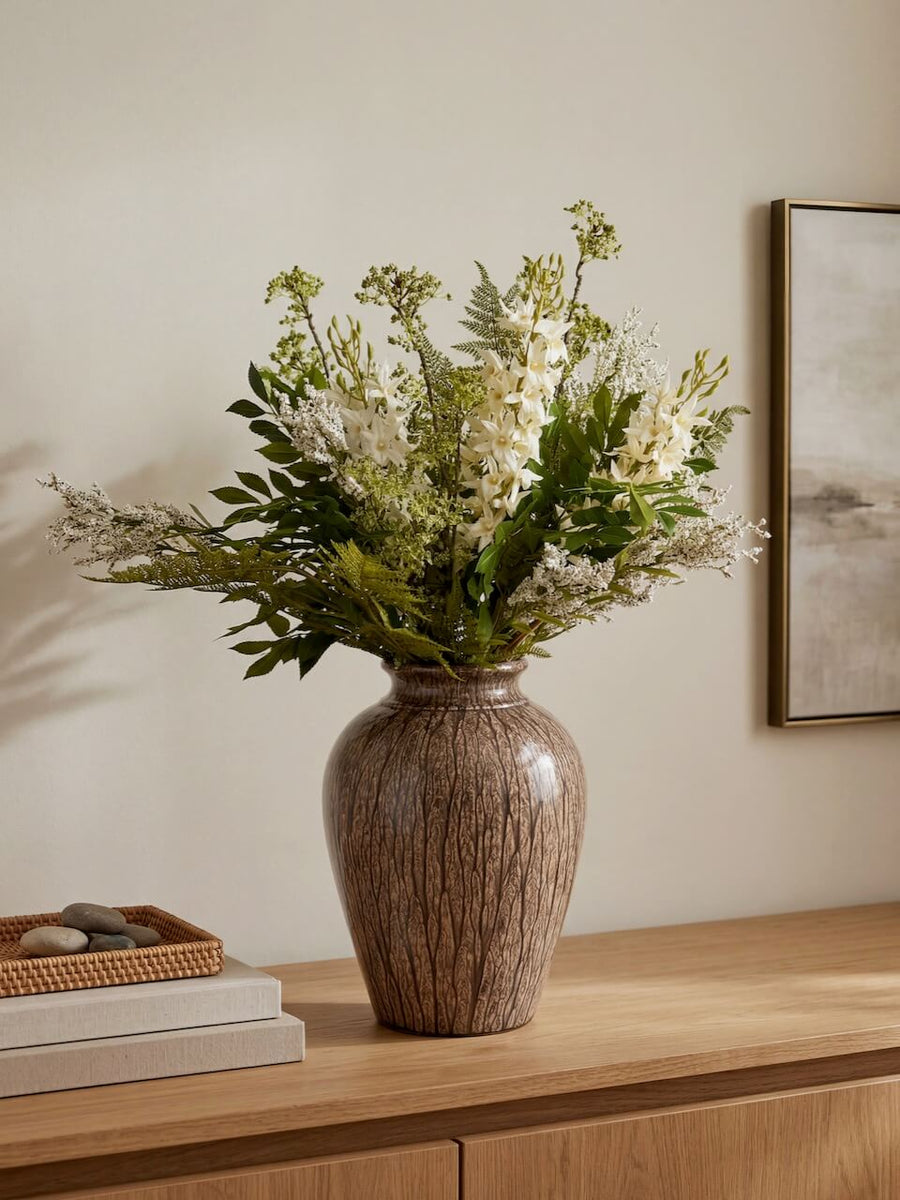 Decorative vase with flowers on a wooden surface against a neutral wall.