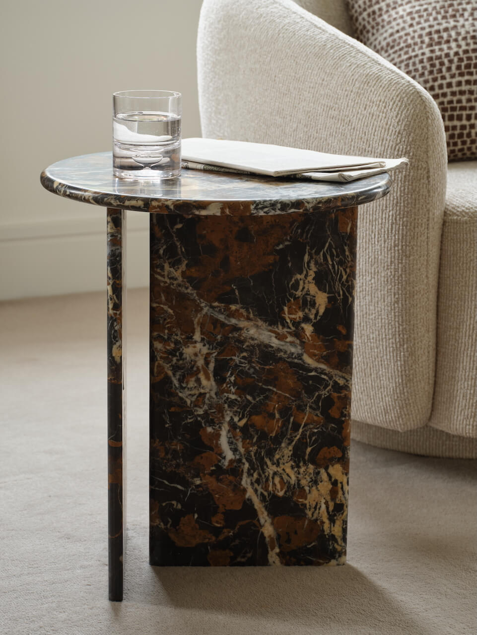 A marble side table with a brown pattern, featuring a glass of water and a remote on top, placed next to a chair in a room with a neutral color scheme.