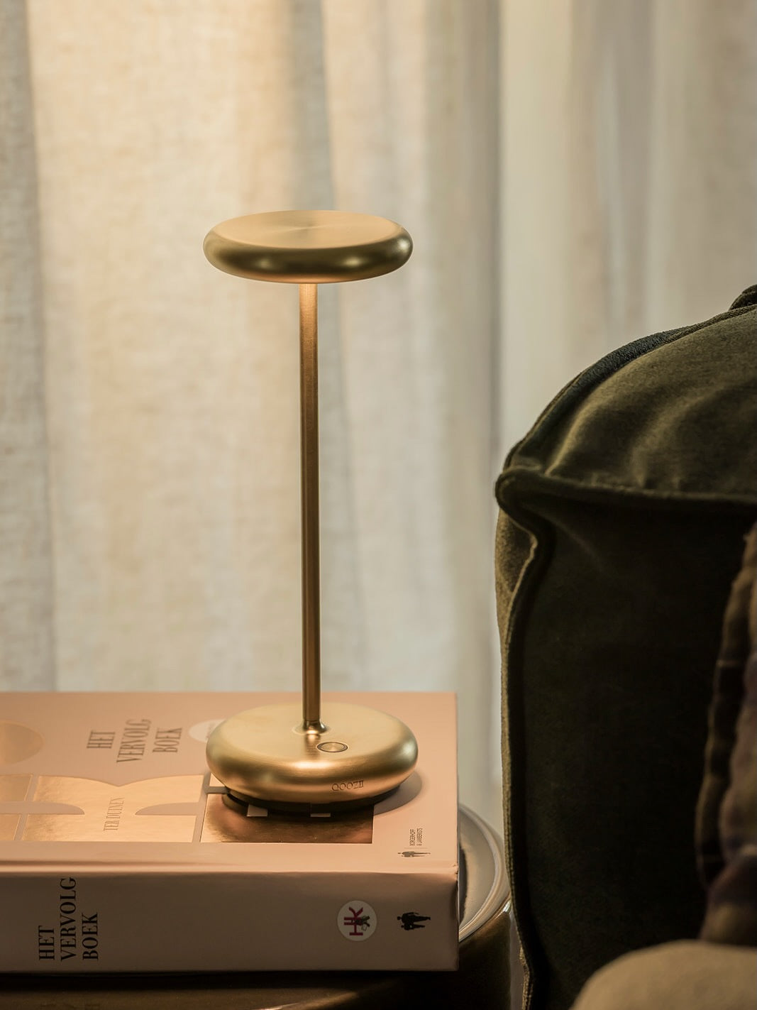 A modern, minimalist table lamp with a brushed brass finish, featuring a circular flat top and a matching circular base, shown resting on a book next to a dark green armchair in a cozy indoor setting.
