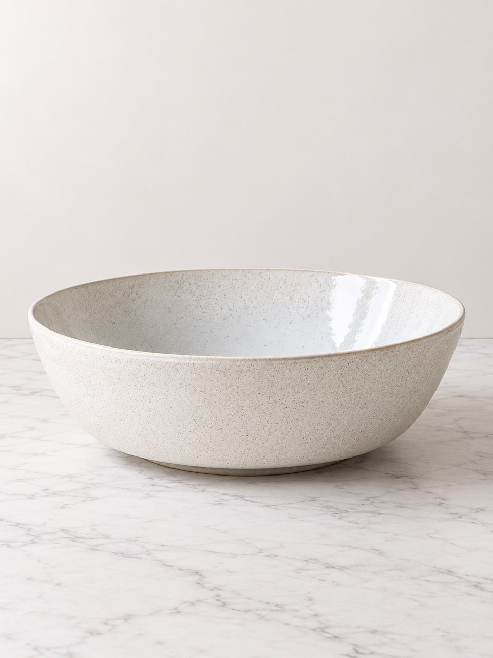 A large round ceramic serving bowl with a speckled off-white exterior and a glossy pale blue interior, set on a white marble surface against a neutral light gray background.