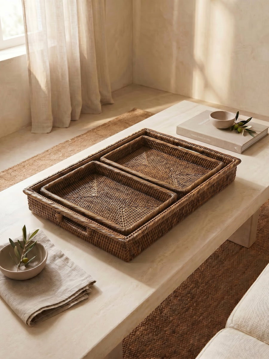 Set of three antique brown handwoven rattan trays styled on a light stone coffee table in a warm neutral living room with linen textiles and soft natural light.