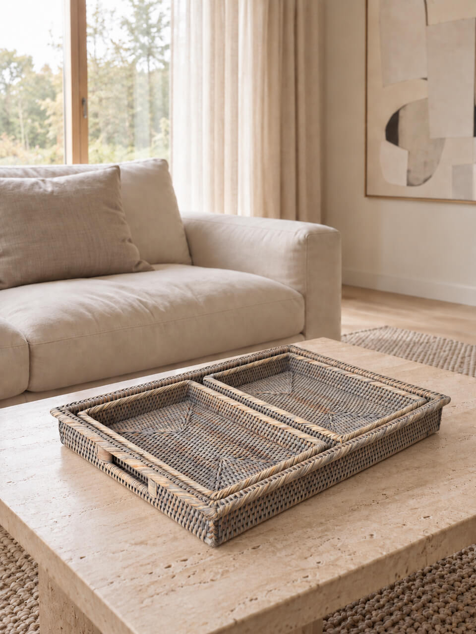 A rectangular woven rattan tray with two divided compartments sits on a light stone coffee table in a modern living room setting with a beige sofa and neutral-toned walls.