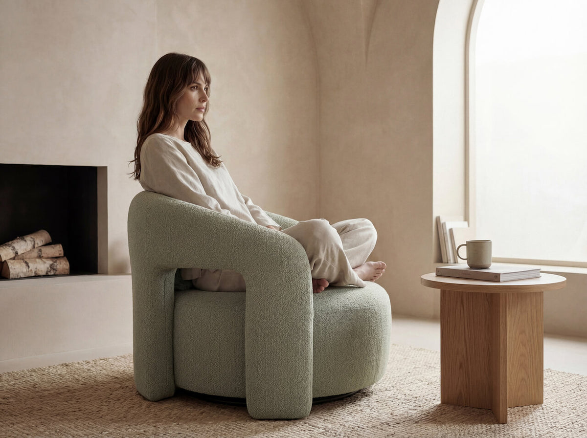 A woman sits cross-legged in a modern, rounded sage green armchair with a bouclé texture in a warm, minimalist room with beige walls, a fireplace, and a small wooden side table.