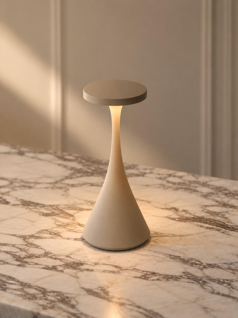 A modern table lamp with a tapered beige body and a circular top, shown lit on a marble surface against a neutral paneled wall.