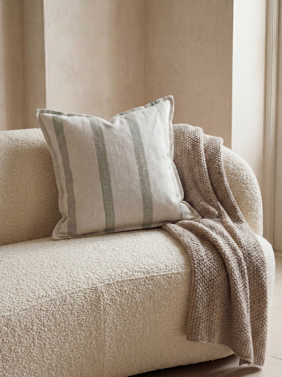 Luxury sage green striped cotton linen cushion cover styled on a textured boucle sofa with a soft knitted throw, creating a warm and relaxed contemporary interior setting.