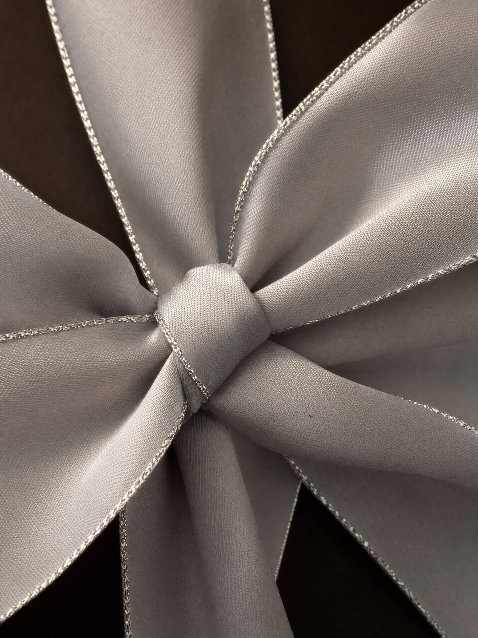 A close-up of a light gray satin ribbon tied into a bow, showing its smooth texture and delicate silver edge trim against a dark background.