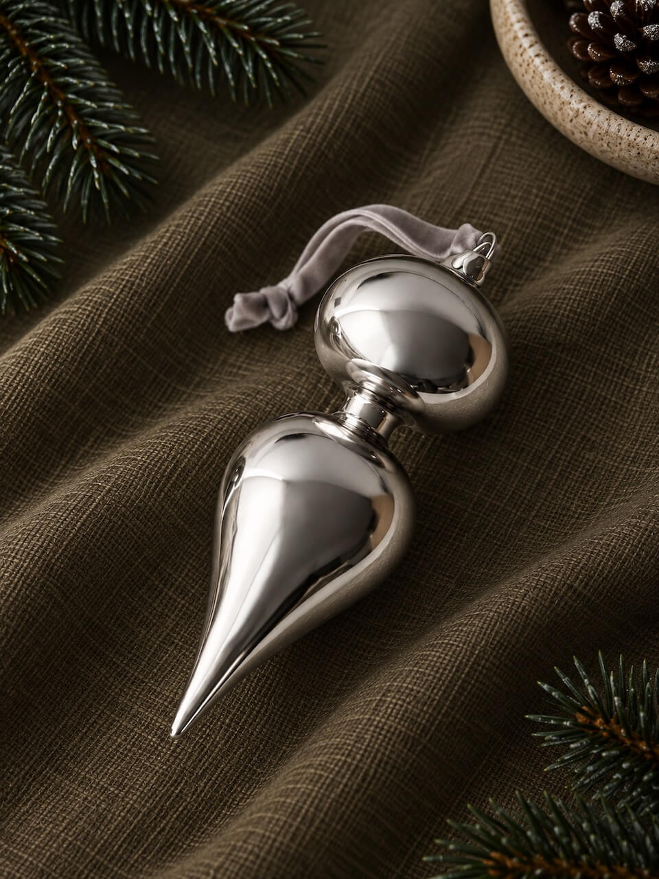A polished silver metallic double-bulb ornament with a small gray fabric loop, resting on a textured dark brown fabric surface with pine branches and a pinecone as props.