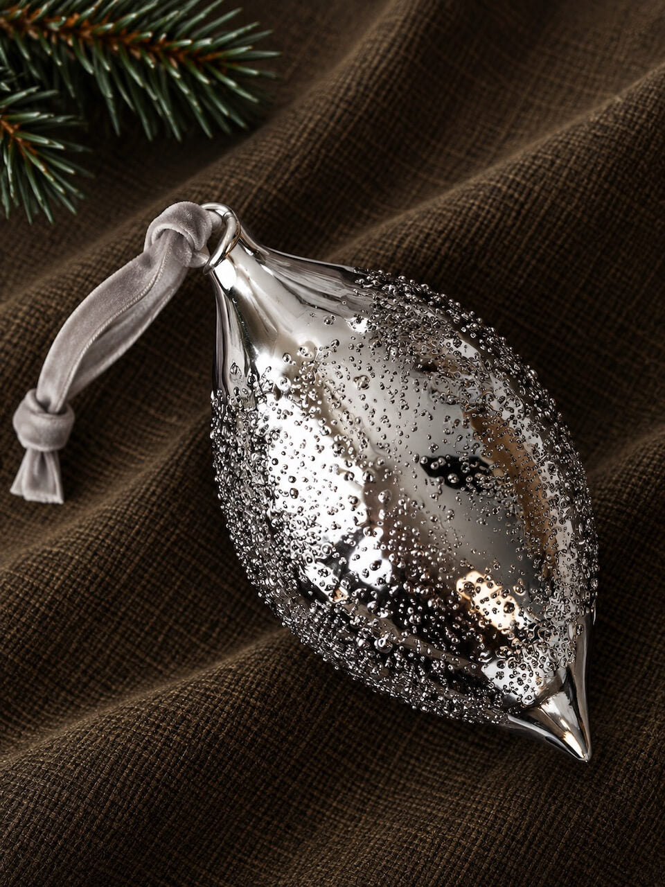A metallic silver Christmas ornament shaped like a teardrop or fish, covered in textured glitter and a small gray ribbon loop, resting on a dark brown fabric background with pine needles in the corner.