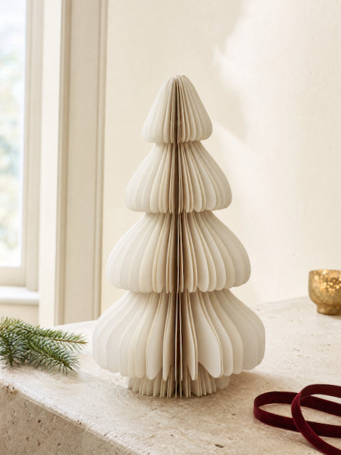 A cream-colored, accordion-style paper Christmas tree stands on a light stone surface in a bright indoor setting. A small green pine sprig rests on the left, and a gold bowl and dark red ribbon are visible in the background.